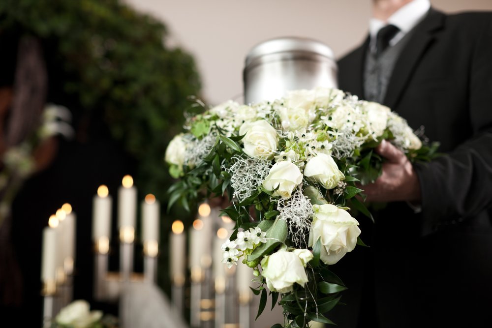 Kegyeleti dolgozó fémből készült urnát tart a kezében melyet körben fehér virágokból készült koszorú övez